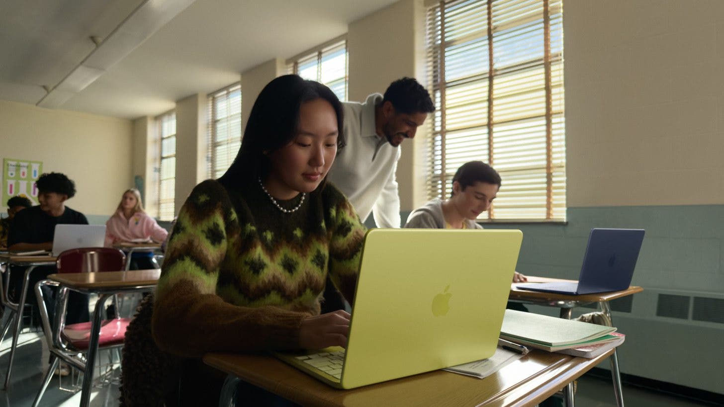 an image of a young woman in a classroom setting using a citrus colored macbook neo at her desk