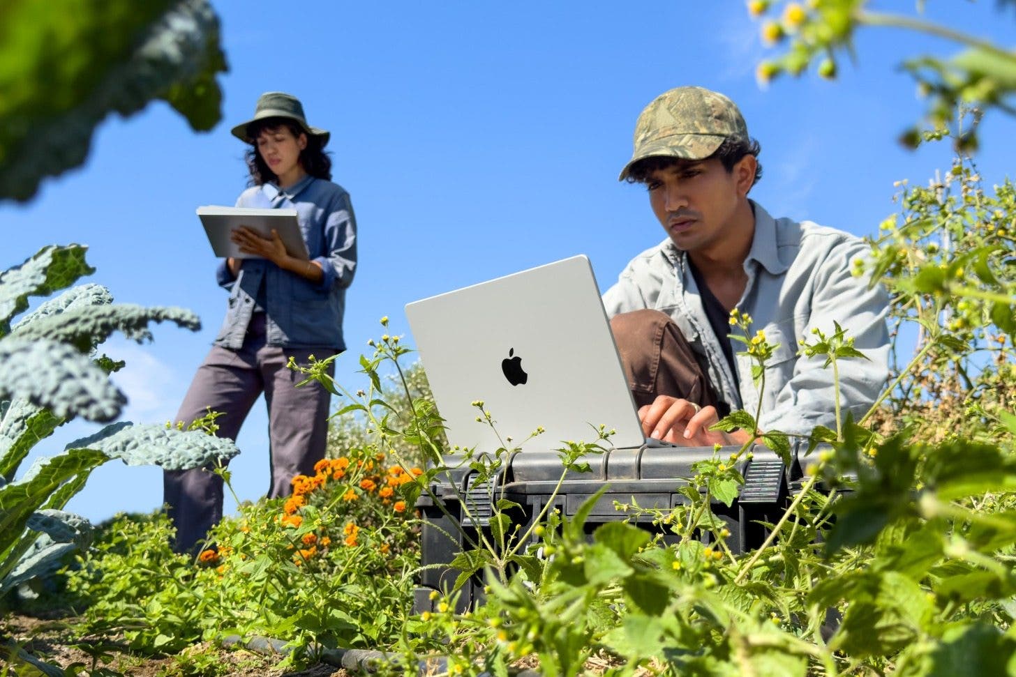 an image of a man and a woman outdoors. the man is sitting at a makeshift desk using a macbook pro